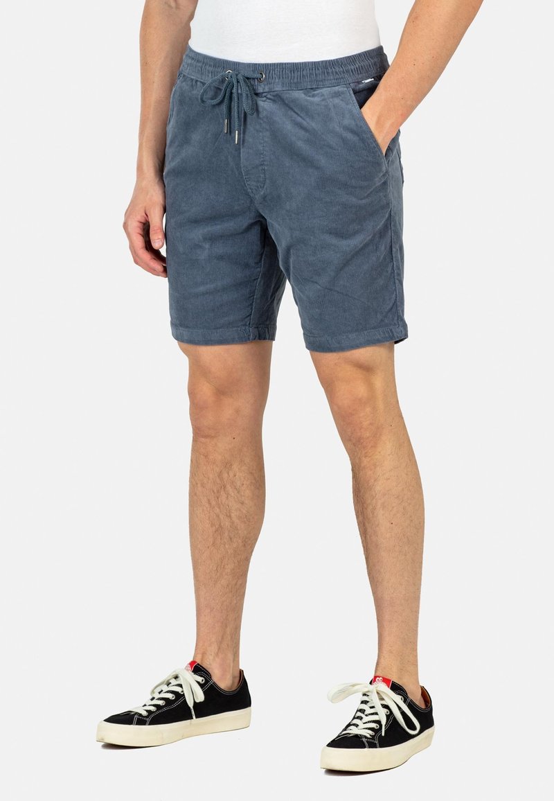 Pantalones cortos de pana en color azul claro para hombre, con cintura elástica y cordón de ajuste, que cuentan con bolsillos laterales y un corte relajado. Combinados con zapatillas negras.