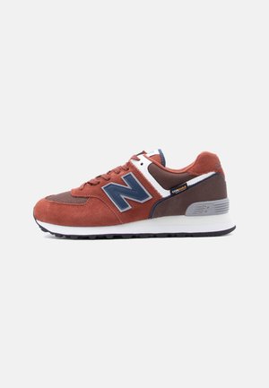 Baskets New Balance marron et rouille avec un logo bleu, des accents blancs et une semelle noire, montrées de profil sur fond blanc.