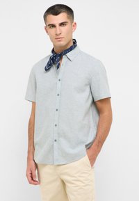 Koszula rozpinana z krótkim rękawem, w jasnoniebieskim kolorze, uszyta z teksturowanej tkaniny, z wysuniętym kołnierzykiem i subtelnymi przeszyciami. Na szyi związany jest granatowy szalik.