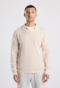 Beige neule, jossa on V-kaula-aukko ja collari, pitkät hihat ja ribatut hihansuut. Päällä vaaleampi paita, yhdistettynä harmaisiin pantsuihin.