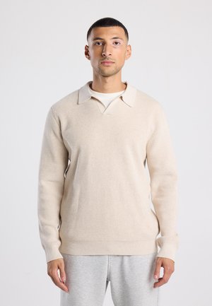 Pull en beige texturé avec un décolleté en V et un col, doté de manches longues et de poignets côtelés. Porté par-dessus une chemise plus claire, associé à un pantalon gris.