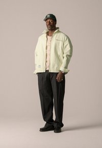 Veste vert clair avec un col montant, poches zippées ; chemise beige en dessous ; pantalon noir ; chaussures noires ; casquette verte avec logo.