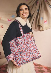 Gėlėtas rankinė su raudonomis ir mėlynomis gėlėmis ant šviesios fono. Reguliuojamos audinio petnešos, blizgus paviršius ir nedidelė etiketės detalė.