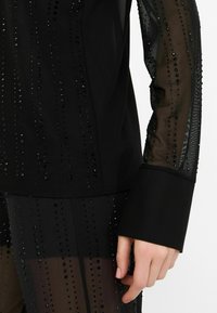 Haut noir avec des manches transparentes, présentant des lignes verticales de petits perles noires. Le tissu est lisse, avec un poignet structuré sur la manche.