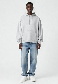 Felpa grigia con cappuccio e tasca a marsupio, vestibilità comoda. Indossata con jeans azzurri chiari e sneakers bianche, su uno sfondo neutro.