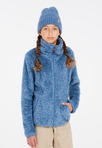 Giacca in pile blu con colletto alto, chiusura a zip e due tasche frontali. Tessuto morbido e peloso; abbinata a un berretto lavorato a maglia blu coordinato.