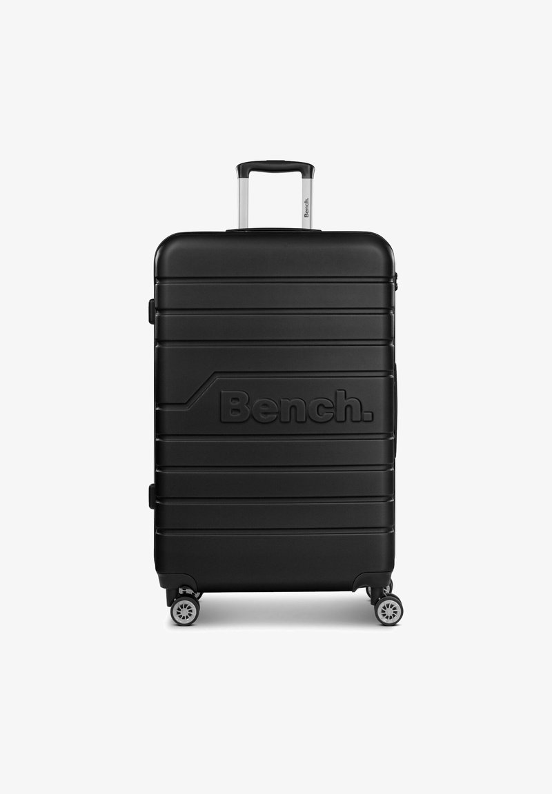 Valigia rigida nera con texture a righe, manico telescopico argentato e quattro ruote girevoli. Presenta un logo Bench. ben visibile sul davanti.