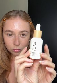 Flaska med serum hudtoning i vit kropp och guldlock. Har en droppare och etikett med varumärkesnamn och produktinformation i svart text.