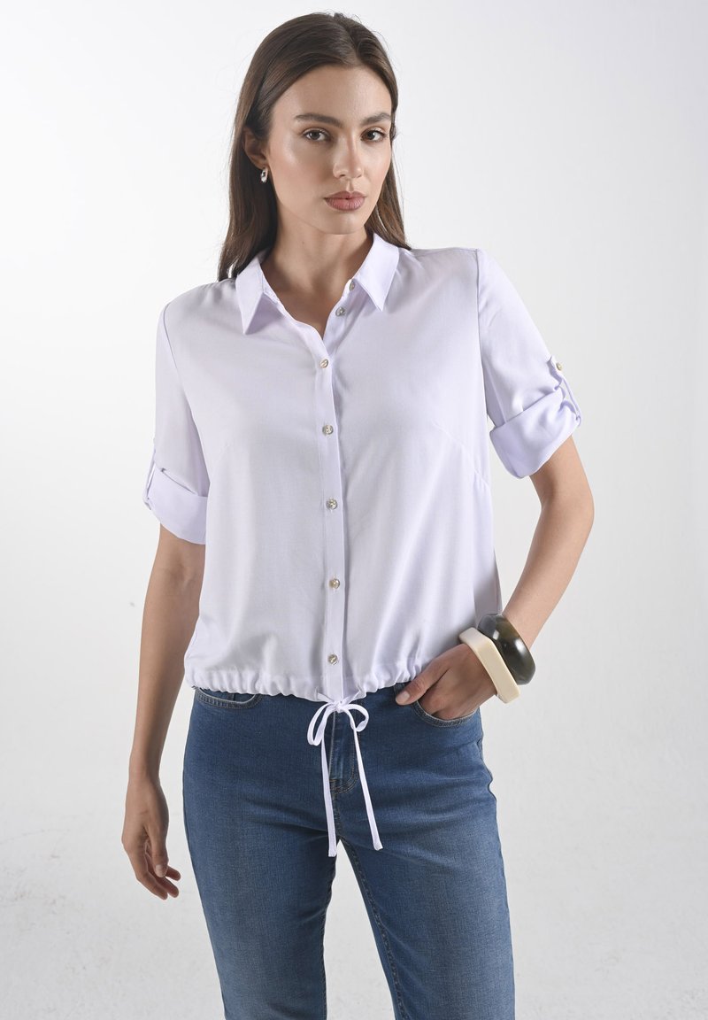 Witte blouse met korte mouwen, knopen, een trekkoord in de taille en opgerolde mouwen. Gladde stof met een subtiele glans. G gedragen met een blauwe spijkerbroek.