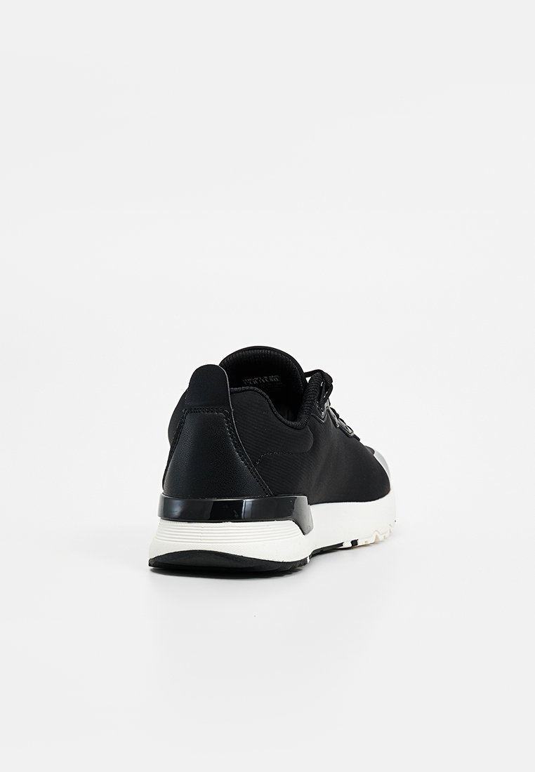 Zapatilla negra con parte superior de sintético suave, entresuela blanca, talón texturizado y sistema de cordones. Presenta un diseño elegante y minimalista con forma redondeada.