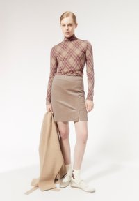 Lange mouwen, bruine geruite coltrui, beige aansluitende rok met een zijsplit, houdt een tan kleurige trui vast, witte sokken en lichte schoenen met banden.