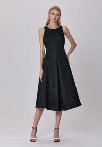Femme debout dans une robe midi sans manches noire avec une texture florale et un collier de perles, portant des sandales à talons noires et blanches.