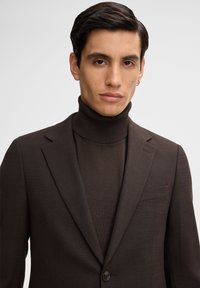 Brauner, maßgeschneiderter Blazer mit einer strukturierten Oberfläche und Reverskragen, kombiniert mit einem passenden Rollkragenpullover. Mit einem einzelnen Knopf und einer Brusttasche.