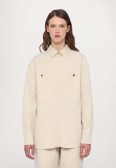 Chemise beige oversize avec un col, deux poches poitrine, des fermetures à boutons et des manches longues, fabriquée à partir d'un tissu doux.