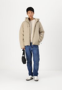 Beige Daunenjacke mit Kapuze, darunter ein leichter Pullover, blaue Cargo-Jeans und schwarze Umhängetasche. Weiße Schuhe mit grauen Akzenten.