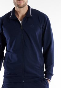 Marineblauwe ritssluitingjas met een hoge kraag, voorzien van een subtiele logo op de borst, contrasterende gestreepte accenten op de kraag en zijzakken.