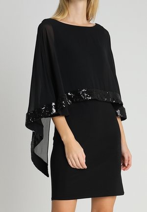 Frau trägt ein schwarzes knielanges Kleid mit einem transparenten Umhang mit Paillettenbesatz, der über die Schultern gelegt ist, vor einfachem Hintergrund.