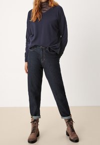 Maglione navy con scollatura a V, jeans dritti scuri e stivali alla caviglia marroni con lacci e suola spessa. Cuffie arrotolate sui jeans.