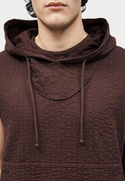 Jong persoon die een mouwloos, textuurrijk donkerbruin hoodiejasje met aantrekkoorden draagt, zichtbaar van schouders tot mond tegen een effen achtergrond.