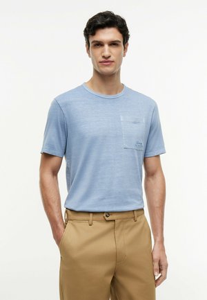 Mann trägt hellblaues T-Shirt mit kurzen Ärmeln und Brusttasche sowie beige Hose, steht mit einer Hand in der Tasche vor weißem Hintergrund.