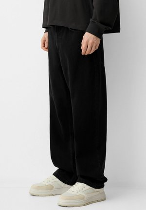 Personne portant un pantalon noir ample et des baskets blanches, debout devant un fond blanc uni, les mains détendues le long du corps.