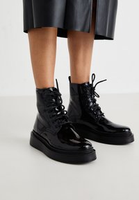 Botines de charol negro con cordones, punta redonda y suela acolchada negra. Textura suave y detalles de costura sutil.