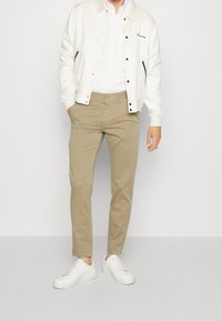 Person som bär beige chinos med vita sneakers, en vit skjorta med knappar och en vit jacka med svarta detaljer och tryckknappar.