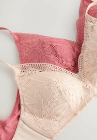 Două sutiene din dantelă, unul roz prăfuit și unul bej, afișate suprapuse pe o suprafață albă, evidențiind modele florale din dantelă și bretele fine.