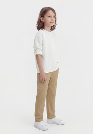 Kind met bruin haar tot op de schouders, gekleed in een wit shirt met lange mouwen, beige broek en witte sneakers, staand tegen een effen achtergrond.