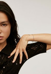 Pulsera de plata con un detalle de símbolo de infinito, llevada en una muñeca. Camisa de encaje negra con cuello y botones decorativos.