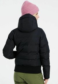 Schwarze Daunenjacke mit tailliertem Design, geripptem Saum und großer Kapuze. Getragen mit einer pinken Strickmütze; Modell zeigt mit dem Rücken zur Kamera.
