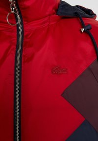 Chaqueta impermeable roja con un acabado texturizado, que presenta un cierre de cremallera plateado, capucha azul marino y formas de acento oscuro. Logotipo de Lacoste en rojo a juego.