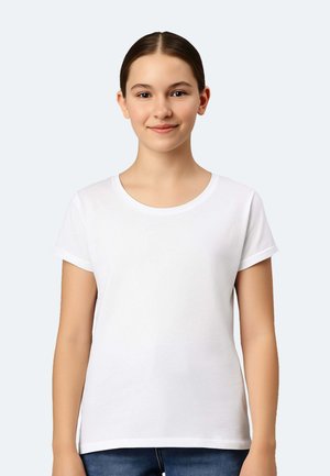 Junge Person mit zurückgebundenem dunklem Haar, trägt ein schlichtes weißes Kurzarmshirt, steht vor hellem Hintergrund und lächelt.