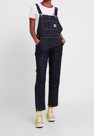 Granatowe ogrodniczki dżinsowe z regulowanymi paskami, pod spodem biała koszula z czerwonym obszyciem oraz żółte trampki z białymi podeszwami.