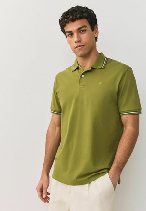 Vīrietis ar cirtainiem matiem, valkājot zaļu polo kreklu ar svītrainu apkakli un aprocēm, stāv pret vienkāršu gaišu fonu.