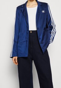 Tmavě modrý sako z saténu se dvěma knoflíky, s výstřihem s klopami a třemi bílými pruhy podél rukávů. Kombinováno s tmavými kalhotami s vysokým pasem.