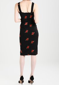 Kobieta w czarnej sukience do kolan z nadrukiem czerwonych róż i czarnych szpilkach, stojąca tyłem do widza.