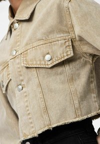 Veste en denim beige cropped avec deux poches poitrine, boutons-pression argentés et ourlet effiloché. Présente un tissu texturé et des lignes verticales.
