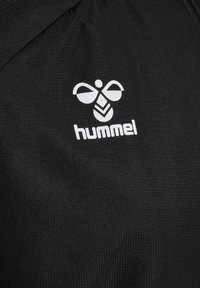 Logo d’abeille hummel blanche et nom de la marque imprimés sur un tissu texturé noir, probablement un vêtement de sport ou un matériau pour veste.