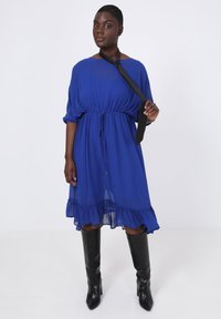 Blauwe chiffon jurk met een trekkoord in de taille en een gerimpelde zoom, gecombineerd met kniehoge zwarte leren laarzen. Lange mouwen en een ontspannen pasvorm.