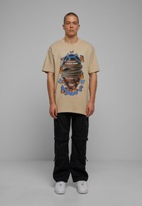 Camiseta de gran tamaño en color beige con estampado gráfico que incluye una cara, mariposas y texto. Combinada con pantalones cargo negros y zapatillas blancas.