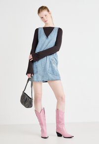 Denimi overall-mekko, jossa on V-pääntie, kaksi etutaskua ja vetoketjusomistus. Vaaleanpunaiset cowboy-saappaat, joissa on pieni korko. Musta pitkähiha顶部.