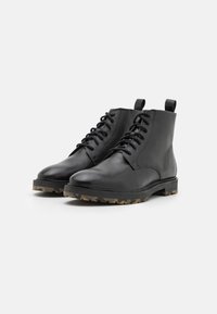 Botas de tobillo de cuero negro con diseño de cordones, tira en la parte trasera y suela de goma resistente con un patrón de camuflaje.