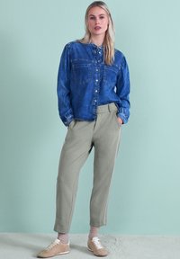 Chemise en denim bleue avec deux poches poitrine, associée à un pantalon de survêtement gris clair fuselé avec des bandes blanches sur les côtés. Des chaussures beiges complètent le look.