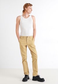 Beige chinos med en lett glans, med knappelukking og lommer, sammen med en hvit tanktop og svarte klumpete sko.