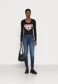 Mujer vistiendo una camiseta de manga larga negra de Guess con un logo floral, vaqueros azules, mocasines negros y sosteniendo un bolso negro frente a un fondo blanco liso.