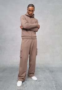 Bruine katoenen rits-hoodie met een capuchon en bijpassende cargobroek, voorzien van zakken en een ontspannen pasvorm. Witte sneakers maken de outfit compleet.