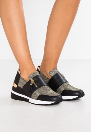 Nogi w czarno-złotych butach typu slip-on z bocznymi zamkami i białymi podeszwami na gładkiej, jasnoszarej podłodze.
