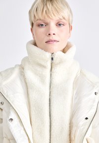 Kermanvärinen puffer-takki, jossa on fleece-ulkopinta ja kiiltävä sisävuori. Varustettu korkealla kaulalla ja vetoketjullisella sulkemisella, mikä tarjoaa lämpöä ja tyyliä.