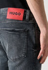 Nahaufnahme einer dunkelgrauen Jeans mit einem roten HUGO-Logo am Bund, die eine Gesäßtasche und die rechte Hand einer Person neben dem Oberschenkel zeigt.